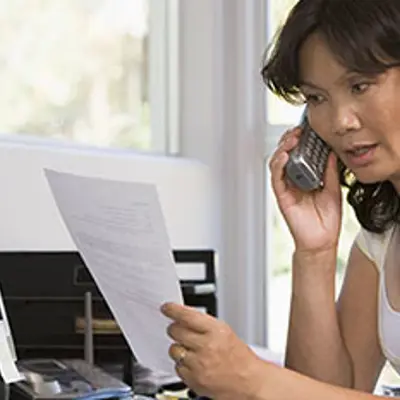 a woman on a phone sitting at a desk in front of a computer holding a piece of paper looking concerned
